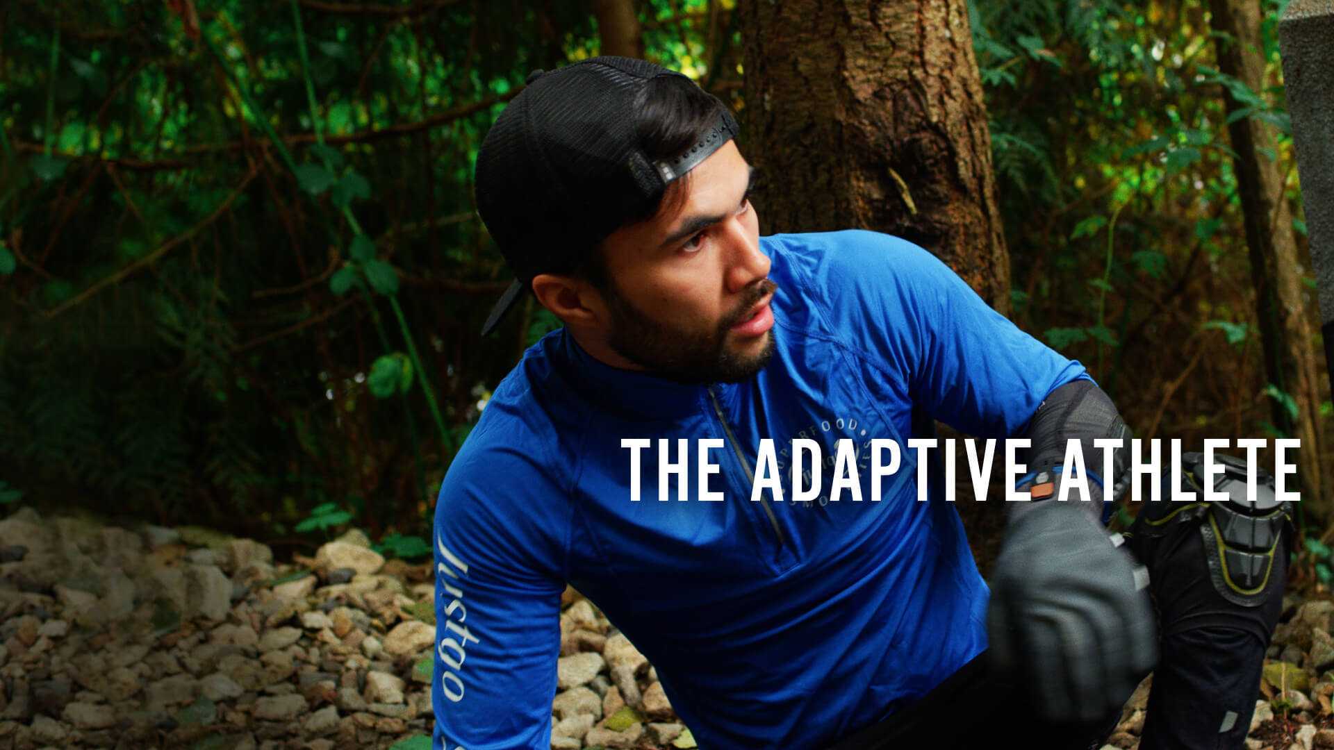 A young man sits on a forest hiking trail while wearing athletic clothing including black gloves and knee protection.