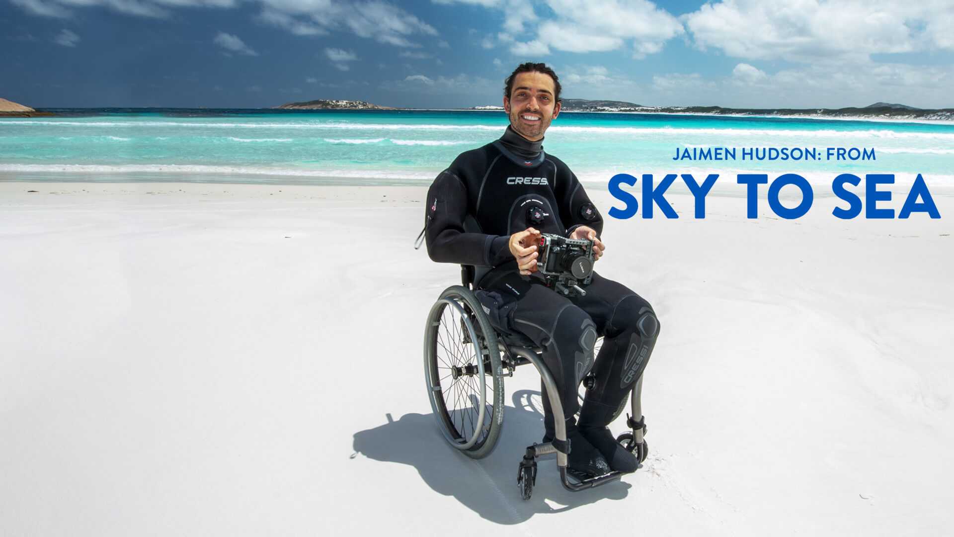 Jaimen Hudson poses on a white-sand beach in his wheelchair wearing a wetsuit. A turquoise sea glistens in the background.