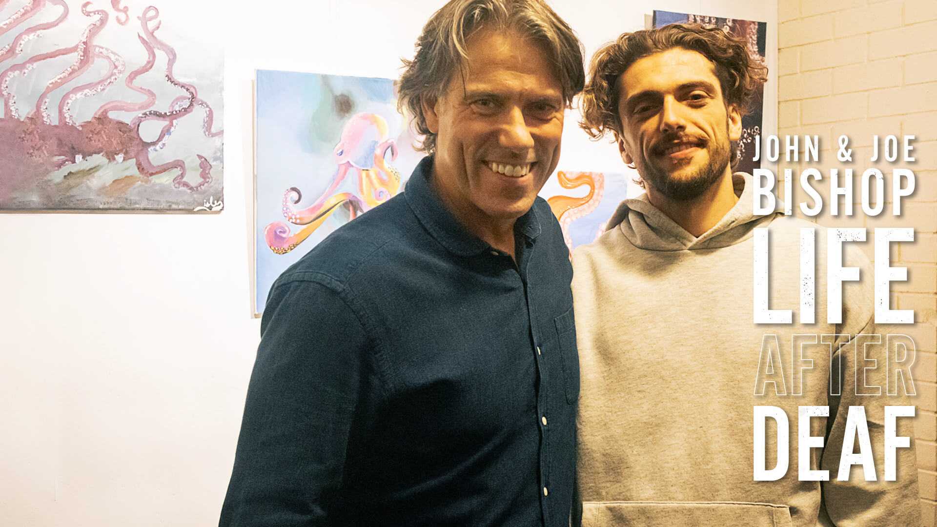 A middle-aged man with a blue jean shirt and younger man with a beige hoodie pose side by side with smiles for the camera.