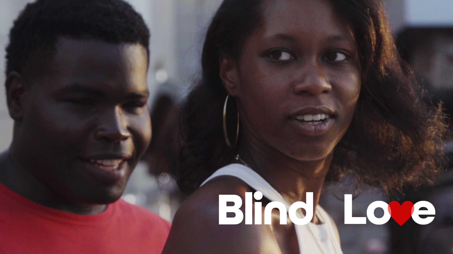 A visually impaired man wearing a red shirt accompanies a woman with a white tank top. The man has a slight smile.